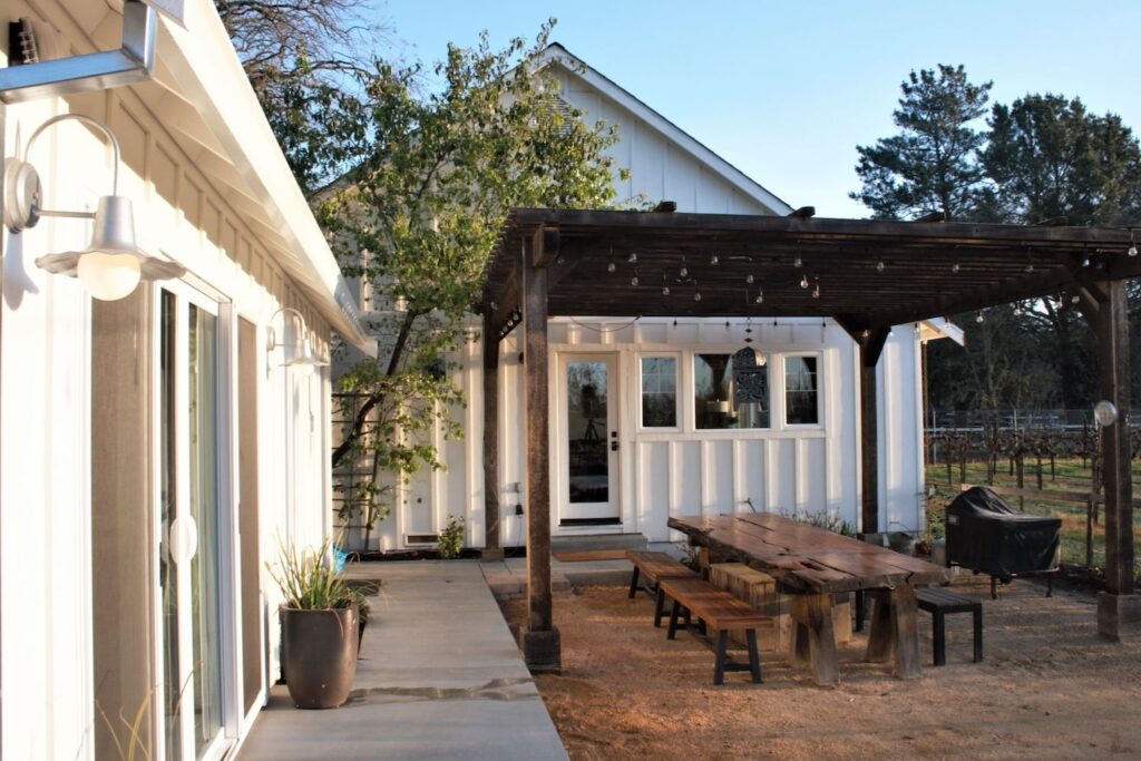 A dark wooden pergola strung with bistro lights covers a picnic table outside a white farmhouse