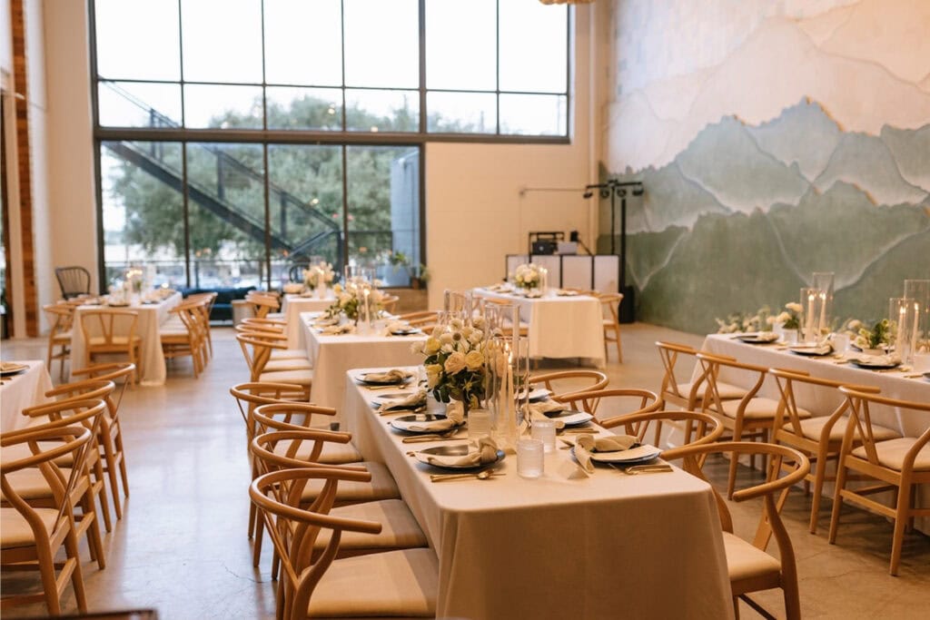 Bright event space with tall windows, a mural of mountains, and simple tables with delicate floral arrangements and modern wooden chairs