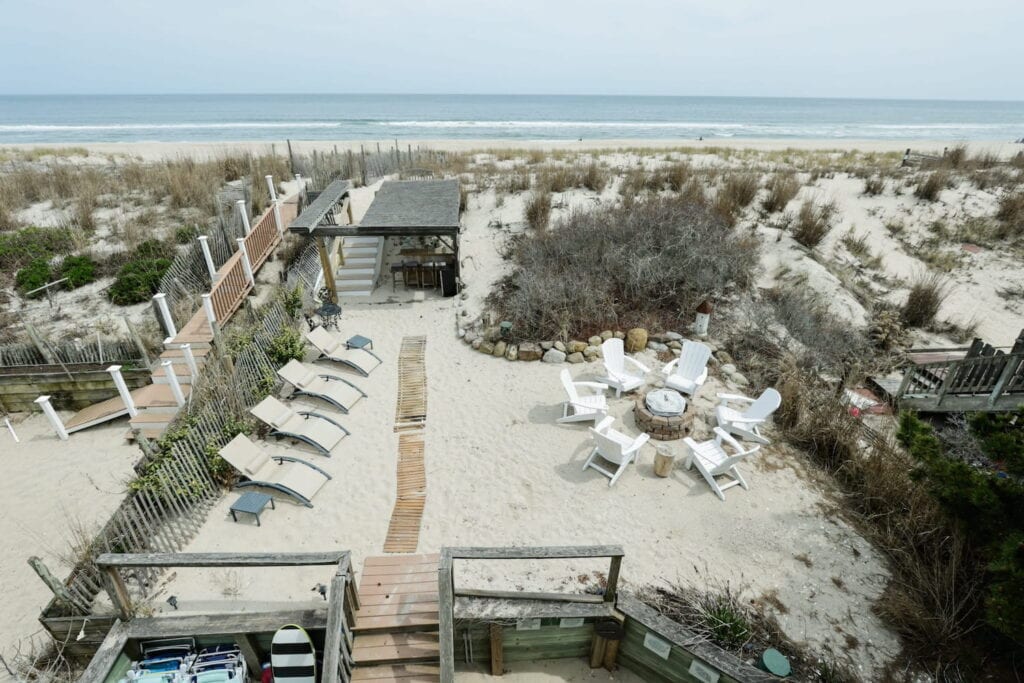 Adirondack chairs and lounge chairs sit in the sand by a pathway to the beach