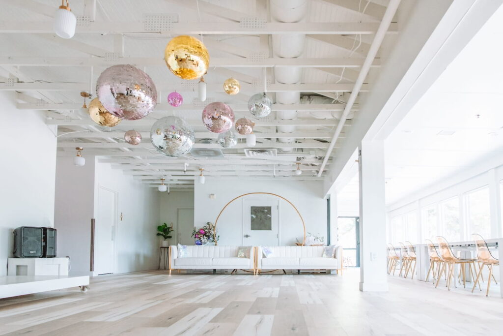 Large, bright event space with white couches, clear dining chairs, and different colored disco balls hanging from ceiling