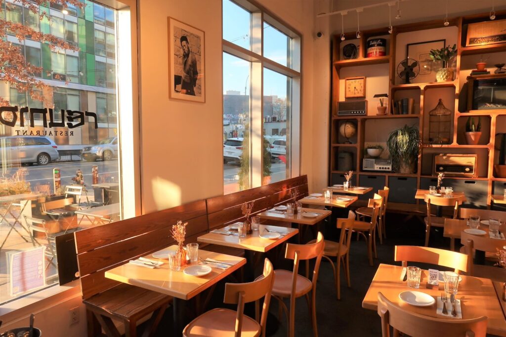 A cozy, soft-lit bistro space is warmed by sunlight from large windows looking out onto a busy street