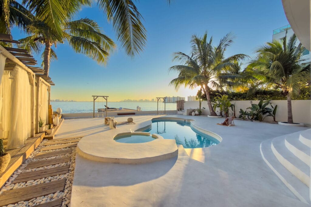 Serene, waterfront backyard pool surrounded by palm trees, light wood cabanas and natural accents