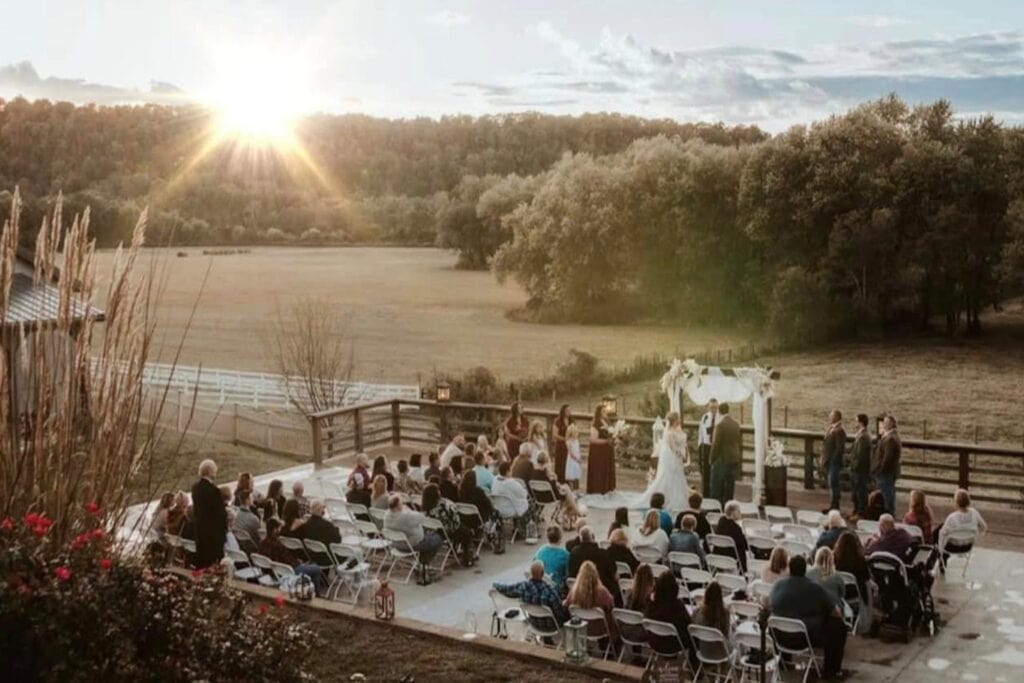 A couple stands at an altar on a patio next to a farm exchanging vows in front of friends and family as the sun sets in the distance