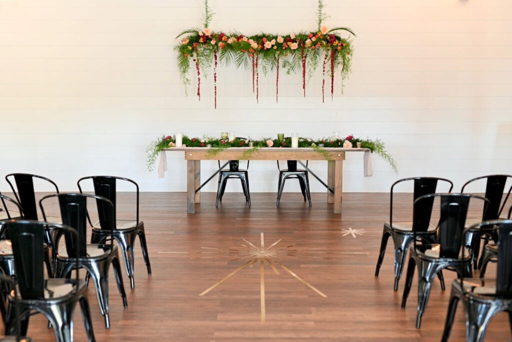 A sweetheart table is set up in an all-white venue space under a canopy of flowers and greenery