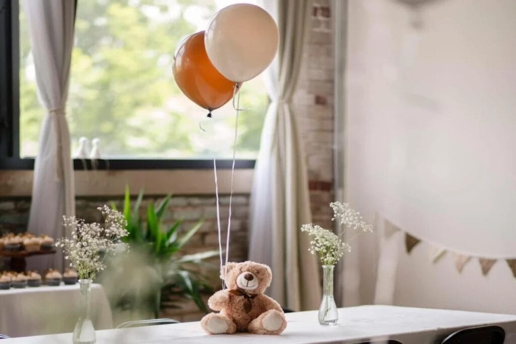 A brown teddy bear sits atop a guest table holding two balloons
