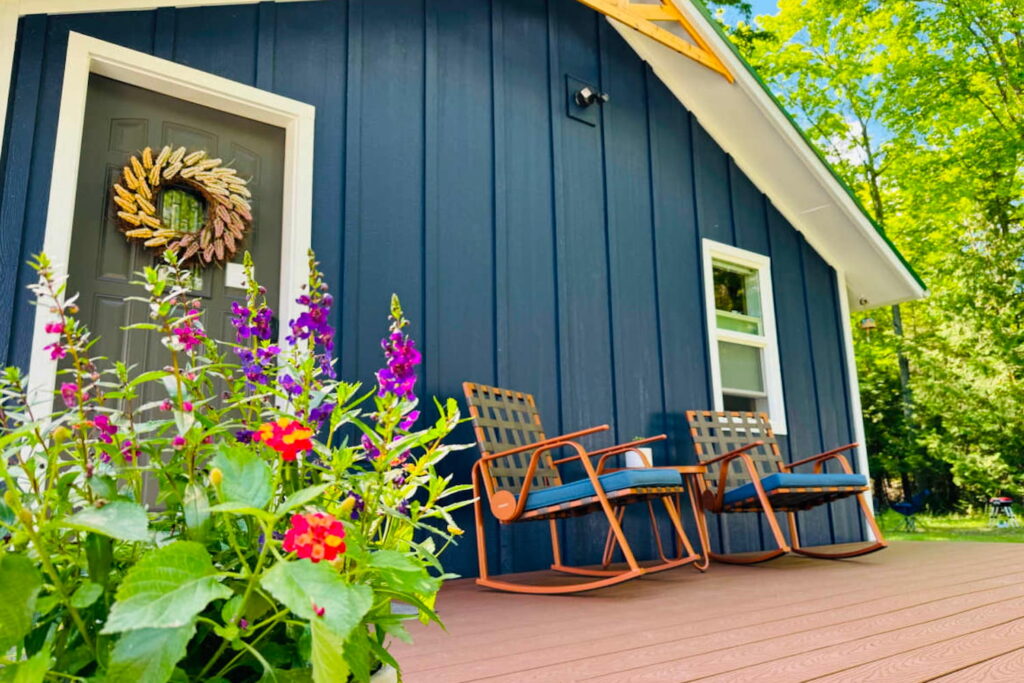 Exterior of a tiny blue cabin in the woods with two rocking chairs on the porch and a bright pot of flowers