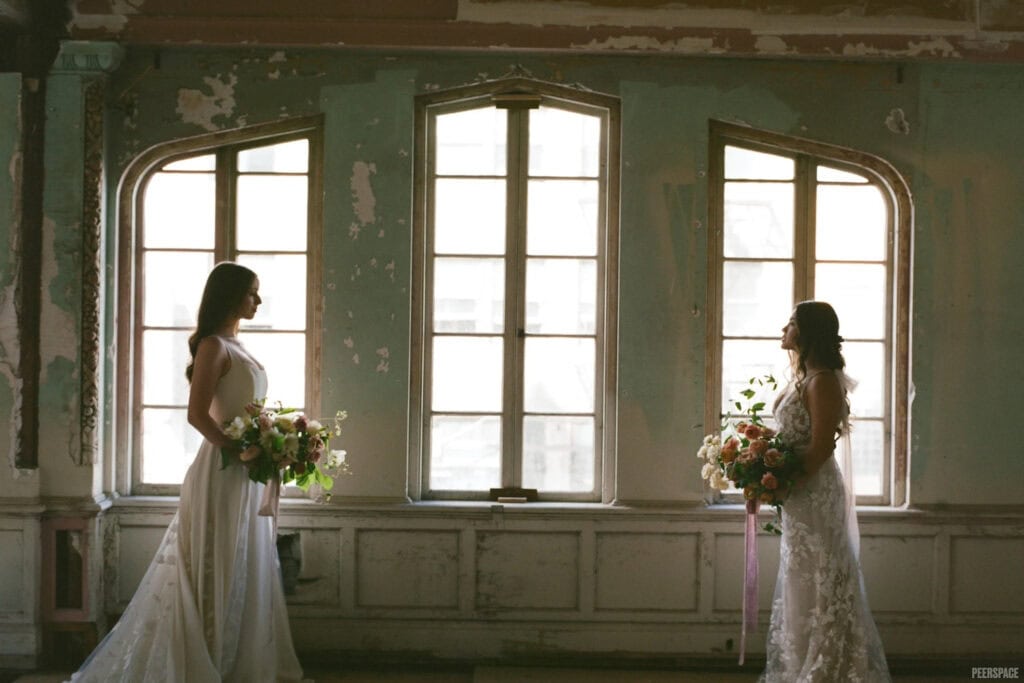 Two brides face each other in front of windows in an historic building