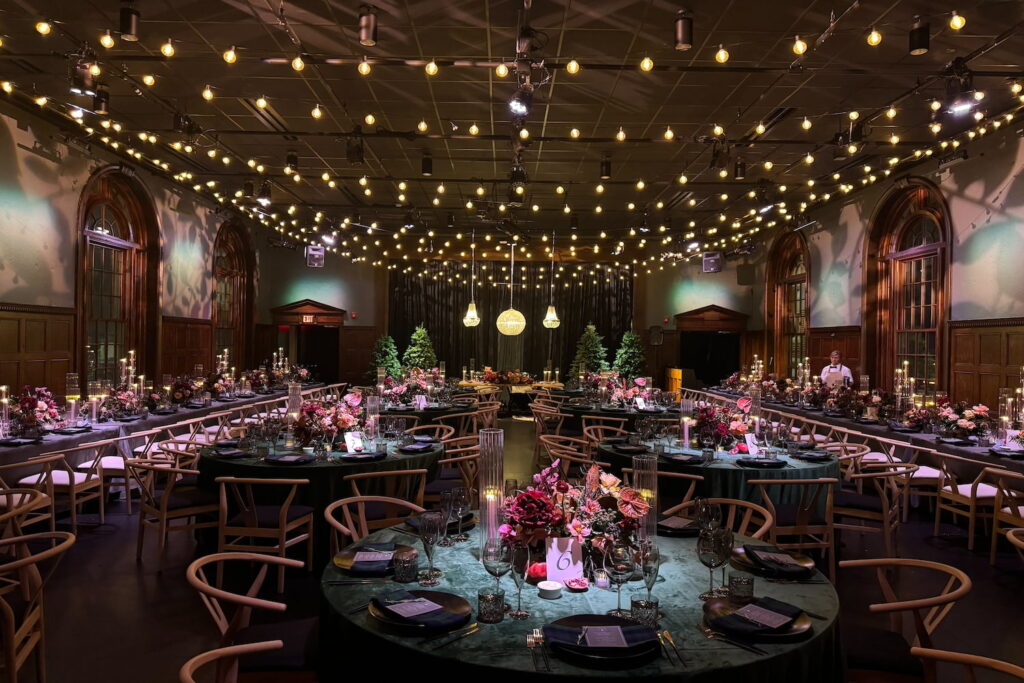 String lights hang above a moody event space decorated in dark green and purple with bold-colored floral arrangements and wood paneling on walls next to huge arched windows and doorways