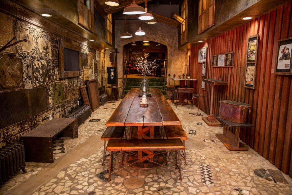 A long wooden table runs down the middle of an urban and industrial event space with wood walls, stone flooring, and dim lighting