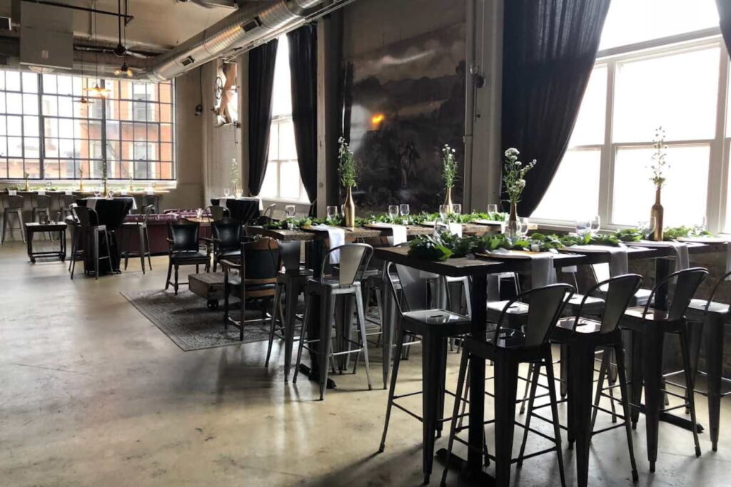 Metal and leather chairs surround wooden tables in an industrial winery space with tall windows and dramatic drapes
