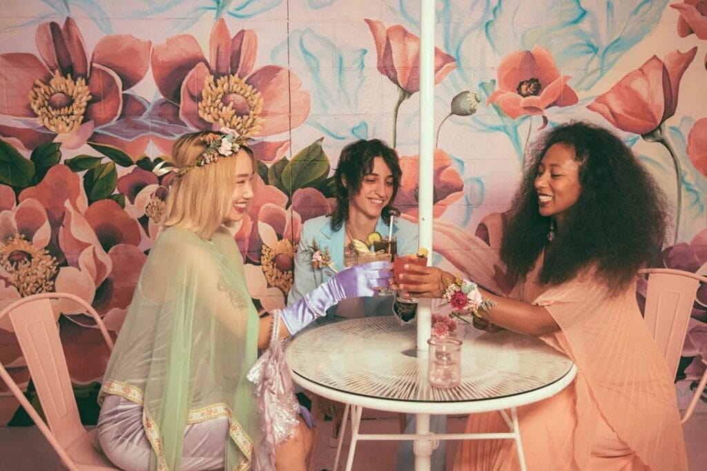Three people toast at a small outdoor table in front of a large, floral mural