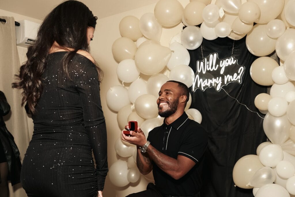 A man is down on one knee proposing to a woman in front of a "will you marry me" sign