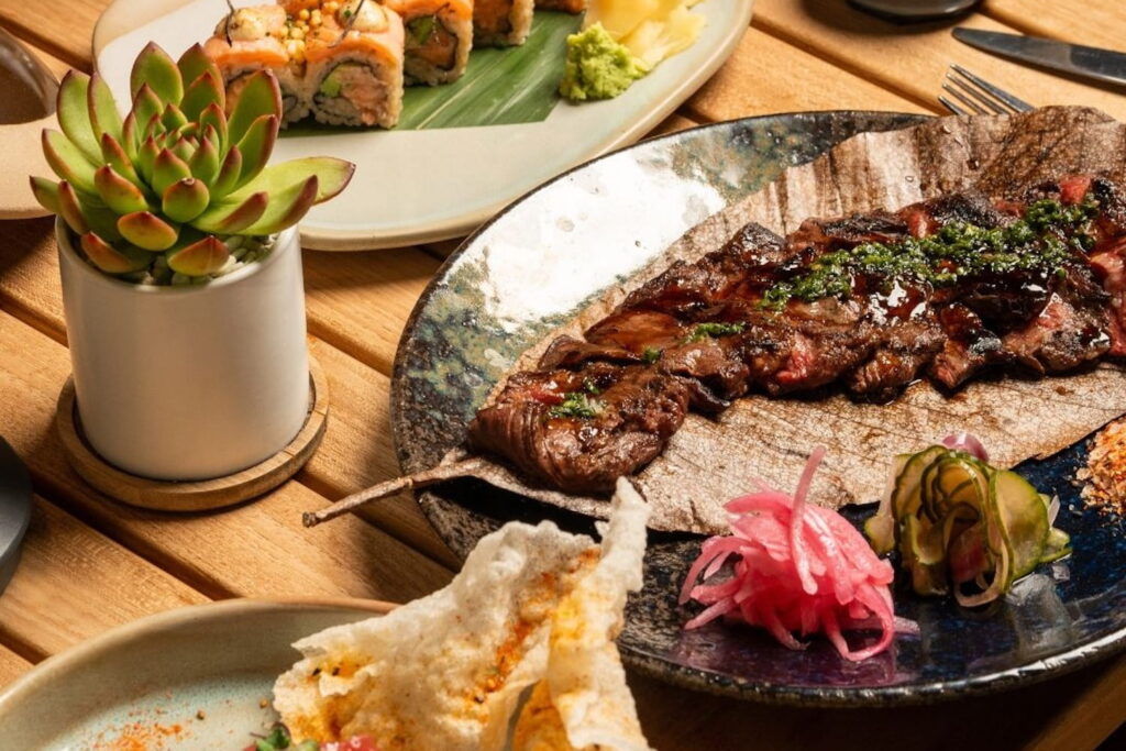 A succulent in a vase stands next to a plate of steak and a sushi roll on a wooden table