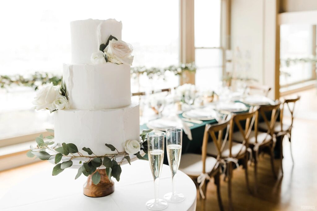 A simple, all-white wedding cat sits in the foreground surrounded by champagne flutes and a brightly-lit window in the background