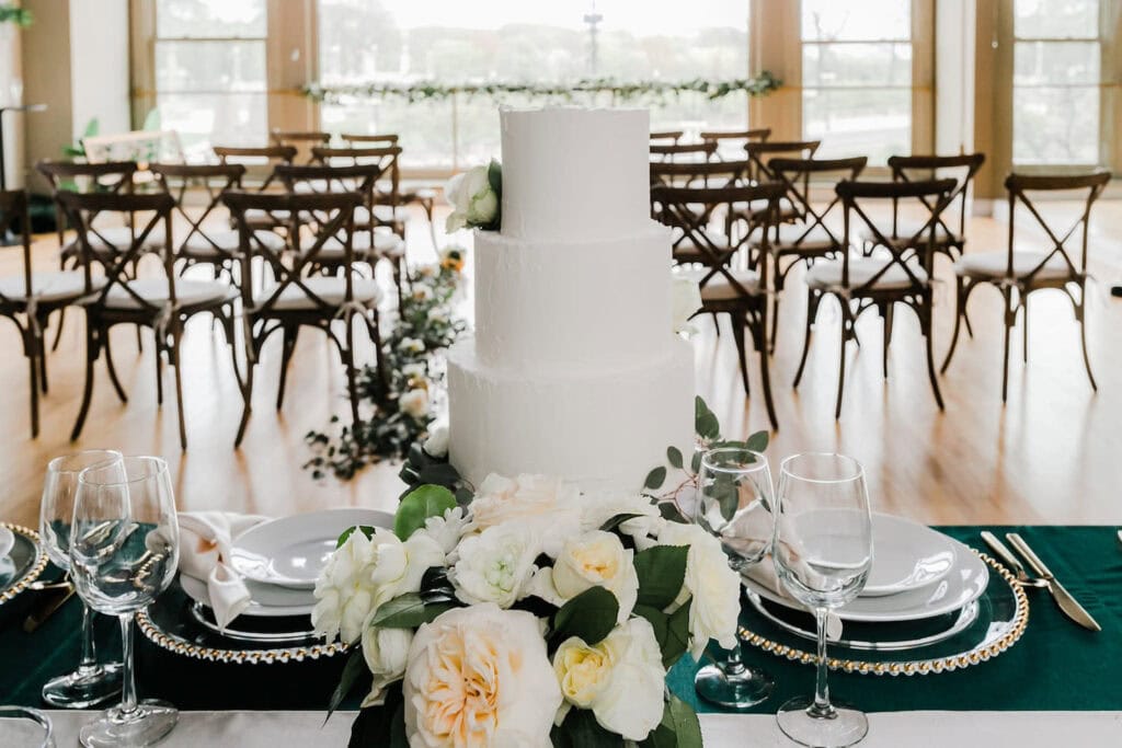 An all-white wedding cake sits on a table with wooden chairs set up for a wedding ceremony in the background facing a window