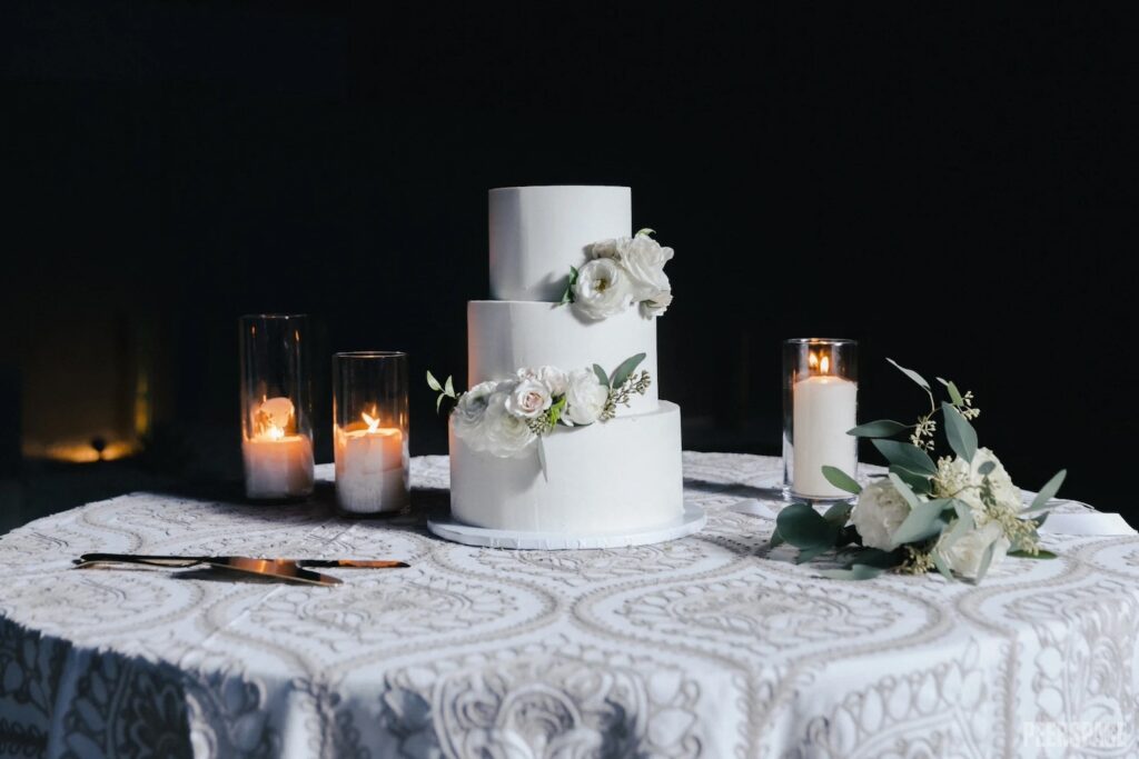 An all-white wedding cake sits on a small table with candles and flowers