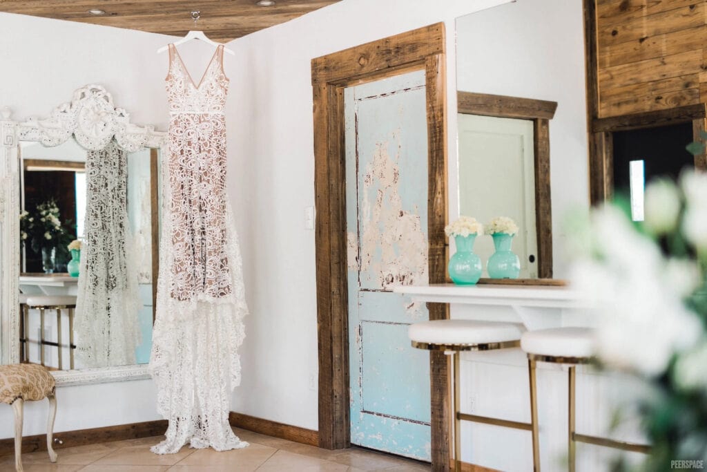 A beautiful lace wedding dress hangs in front of a mirror in a rustic bridal room with barn wood and aqua accents