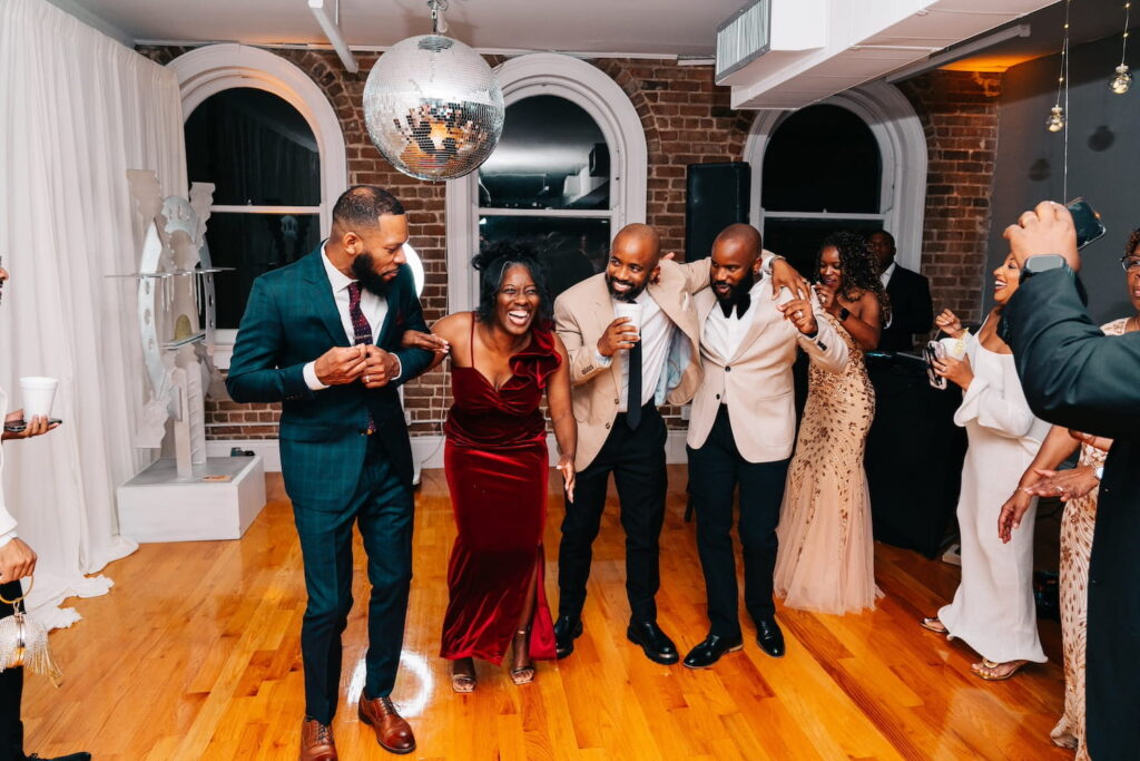 Wedding guests dance on a dance floor beneath a disco ball in an industrial-style reception space with brick walls
