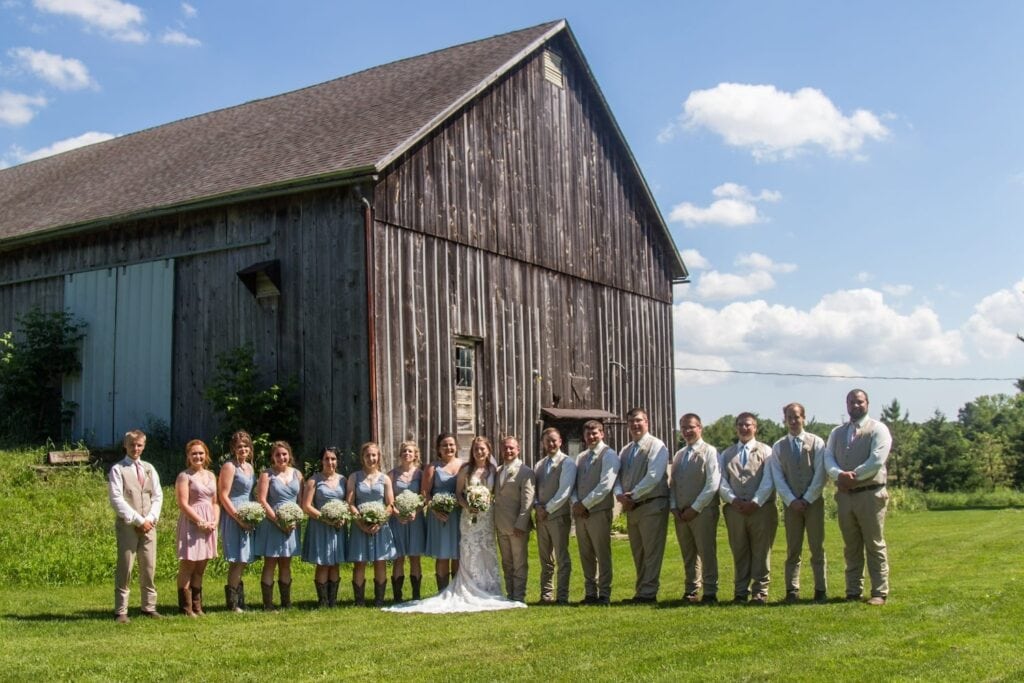 A large wedding party stands beside a bride and groom on a bright, sunny day out front of a rustic barn