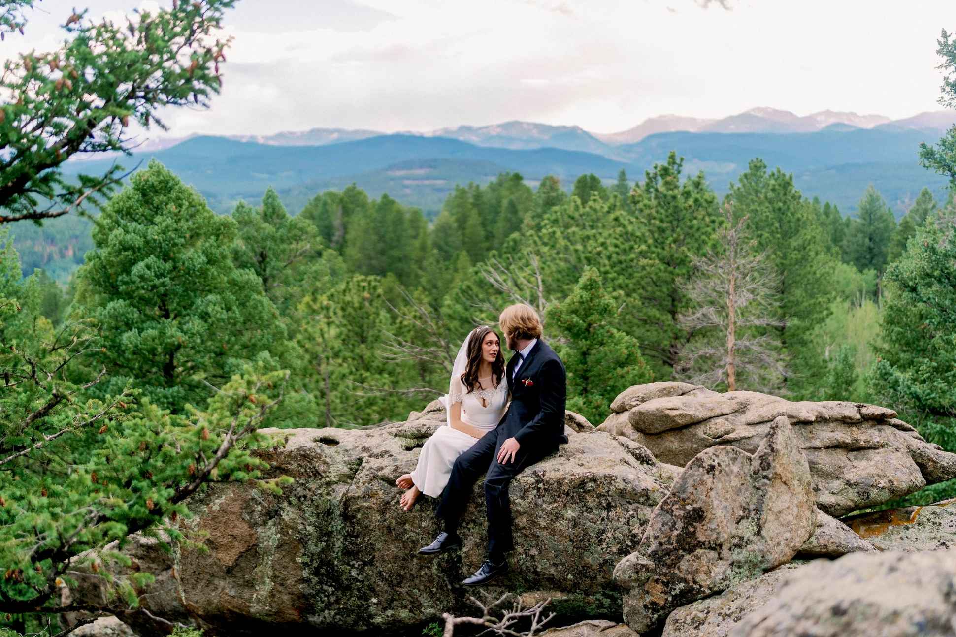 Wedding couple seated on boulders overlooking Alaska forested mountains