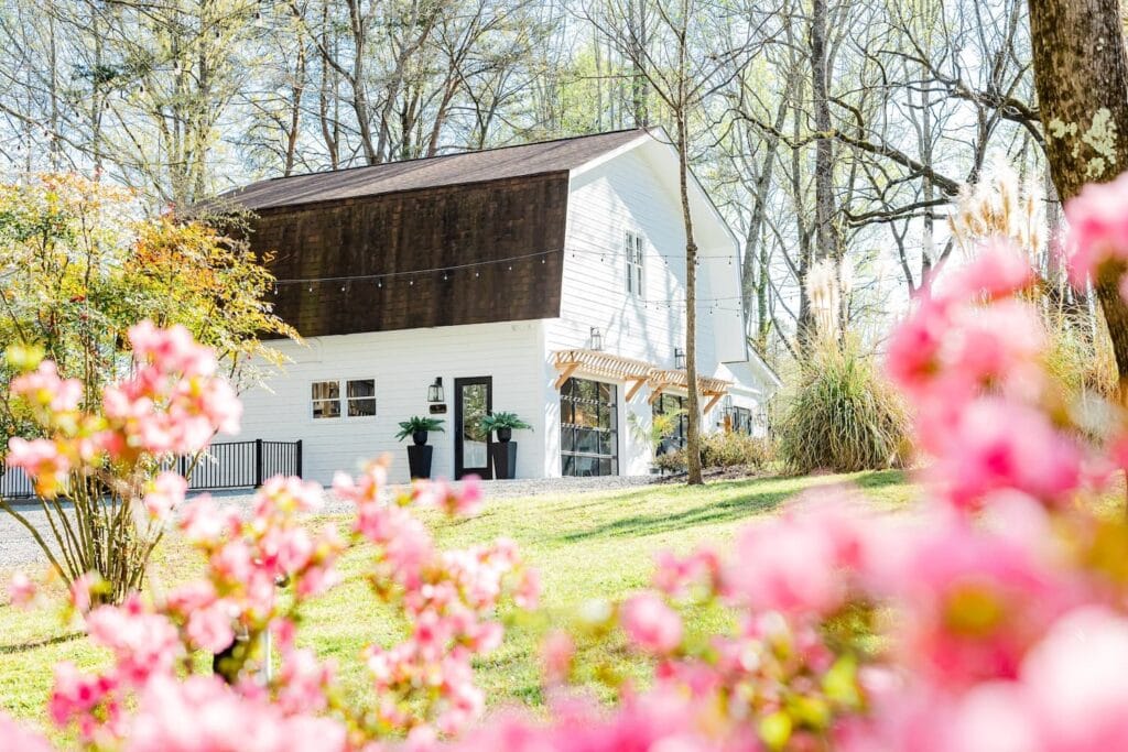 A white barn venue with a brown roof and large garage doors sits in a wooded area surrounded by flowers and string lights