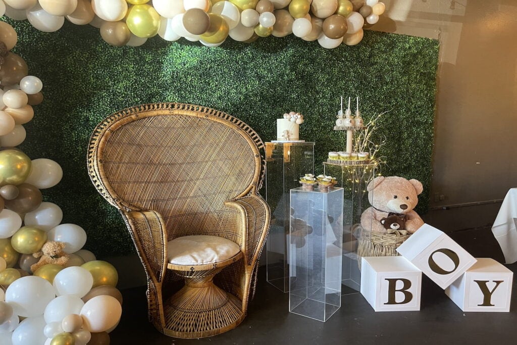 A wicker chair sits in front of a green backdrop with gold and white balloons and baby boy decor