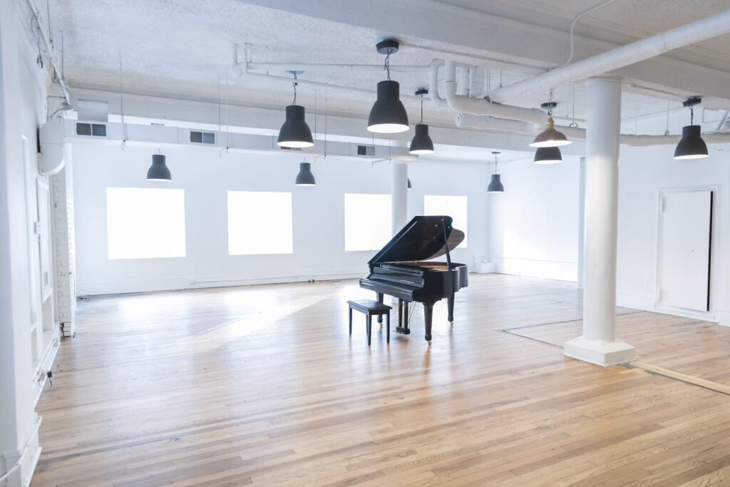 A black, grand piano sits in the middle of an all-white loft space with wood floors and black pendant lighting hanging from the ceiling