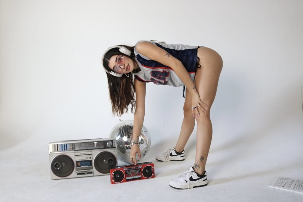 A woman in a sports jersey pushes play on a boombox with another boombox and disco ball in background