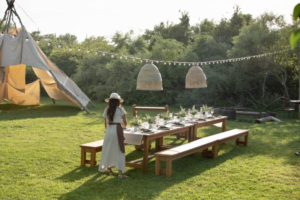 A woman sets an outdoor table under a string of bistro lights and lanterns with a makeshift teepee in the background