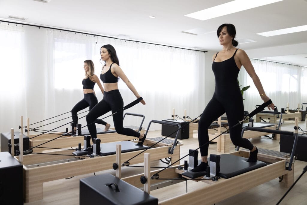 Three women on reformers are doing pilates in a brightly-lit studio.