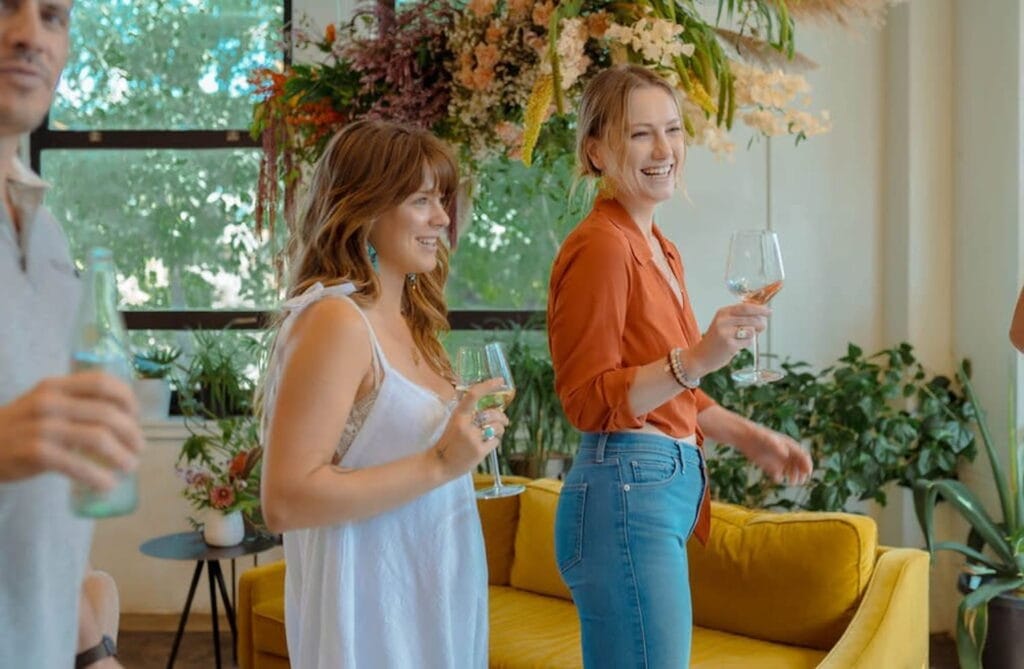Women hold wine glasses while smiling in a cozy, boho space