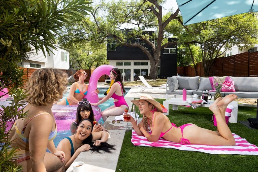 Women in brightly-colored swimsuits sit in and around a pool space at a home surrounded by lush greenery