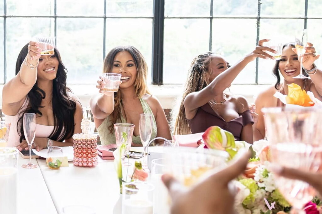 Women seated at a table raising their glasses in a toast with a bright window behind them