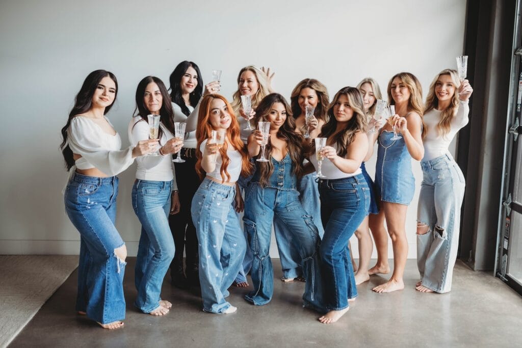 A group of women all dressed in denim and white raise glasses of champagne while smiling at the camera