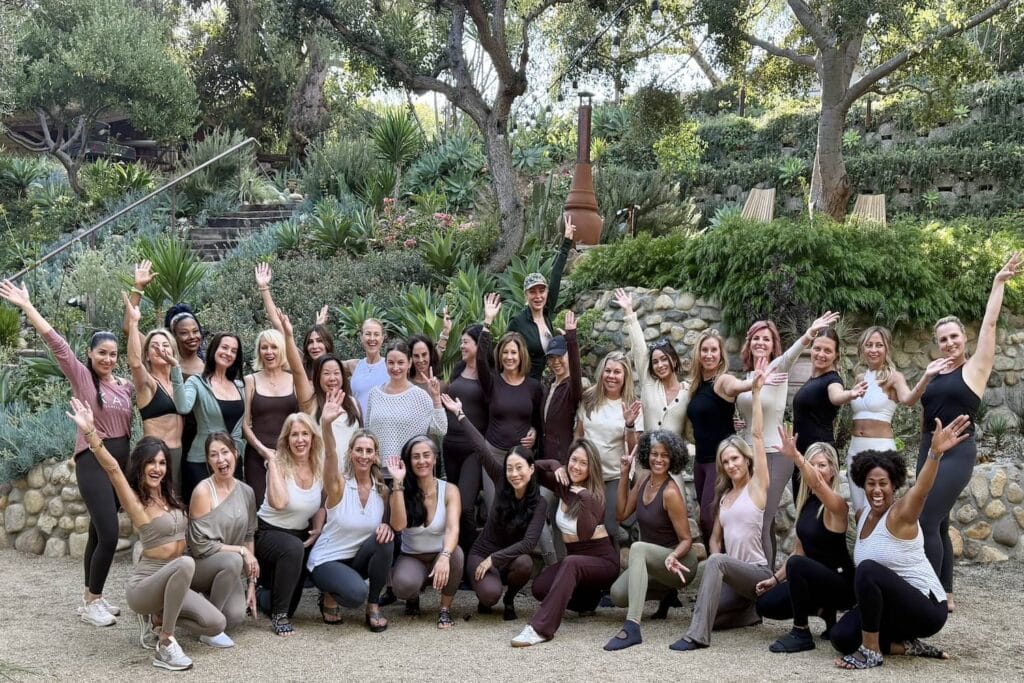 A group of women in workout clothes gather and wave for the camera in front of a lush garden area