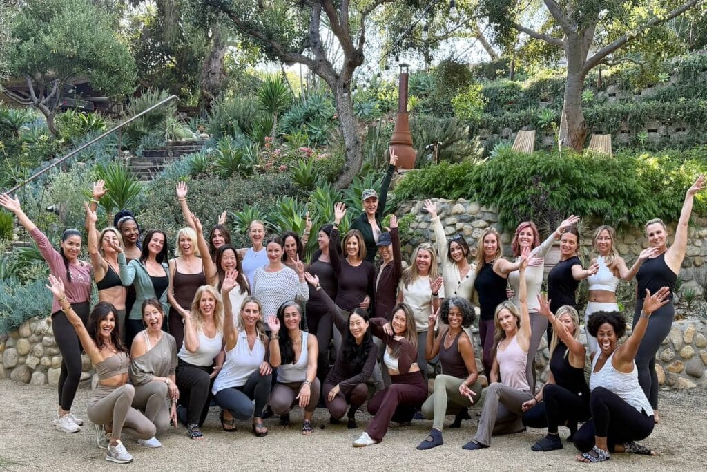 A group of women in workout wear stand together outside waving at the camera near lush greenery