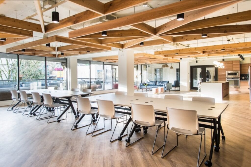An open-plan workspace with long worktables, a kitchen area and a seating area with a large bank of windows and wooden accents on the ceiling
