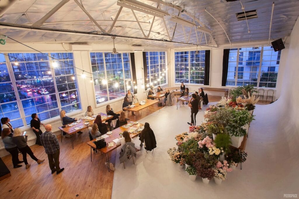 A group gathers from a workshop in a city warehouse space with large windows and twinkly lights.