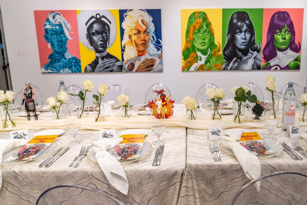 Bright, neon-based portrait art hangs on gallery walls behind a table set with clear ghost chairs and superhero figurines scattered among bud vases holding white roses