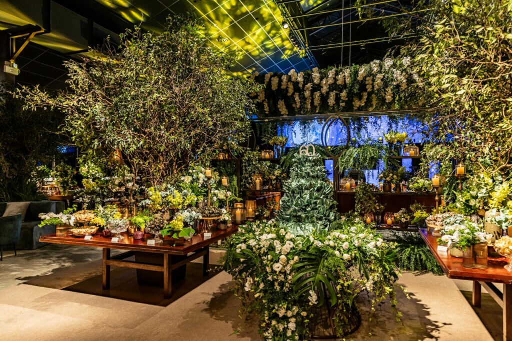 A venue with high ceilings is decorated with lots of plants, flowers, and a 60th birthday cake.