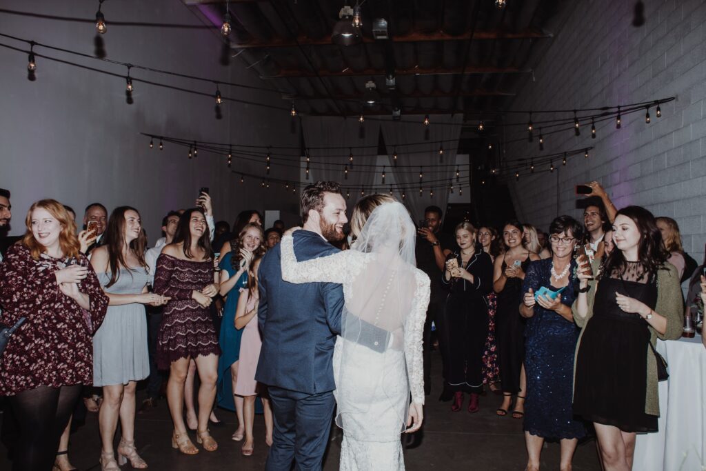Bride and groom dance surrounded by guests at a lively indoor wedding reception venue with string lights overhead.