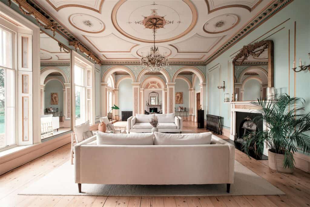 Elegant lounge in a mansion rental with ornate ceilings, chandeliers, arched windows, and plush sofas.