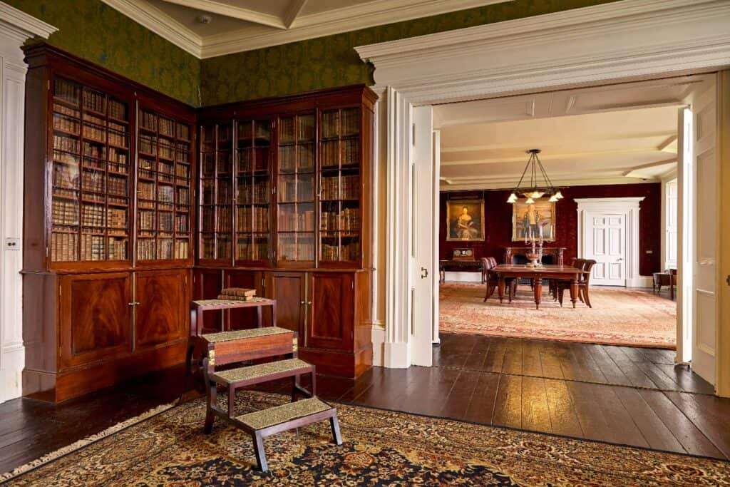 Library with antique bookshelves and classic decor in a grand mansion to rent, with a dining room in the background.