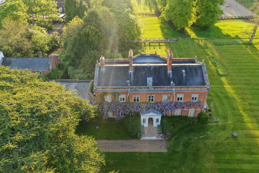 Aerial view of a mansion to rent with extensive green grounds, gardens, and a grand entrance covered in wisteria.