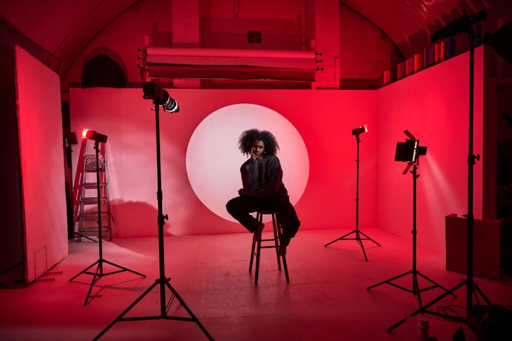 Person posing on a stool in a photo studio with red lighting, spotlights, and a circular white backdrop.