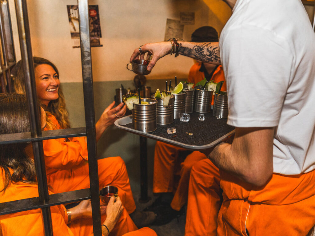 Alcotraz London with guests in orange prison jumpsuits behind bars receiving cocktails served in tin cans by a guard