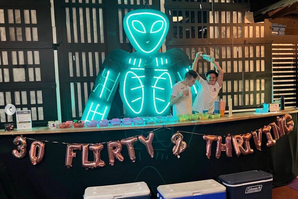 A bar with a neon alien backdrop, 30th birthday balloons and bathenders mixing drinks.