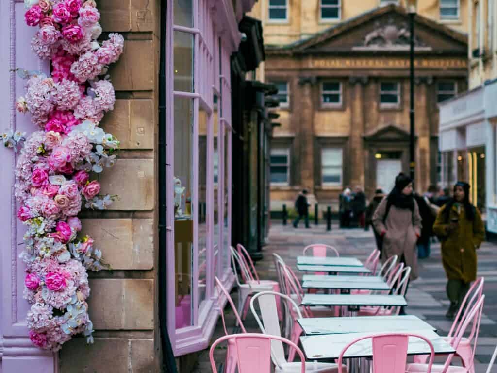 A pink-themed cafe in Bath, UK with flower decoration on the exterior and pink chairs on the street.