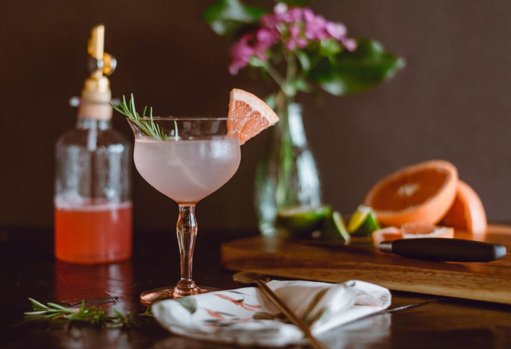A light pink cocktail with grapefruit and rosemary in the foreground and cocktail-making utensils in the background.
