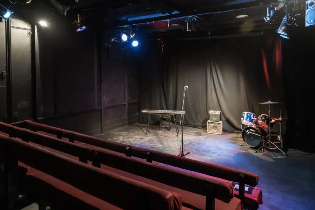 Small performance venue with lit stage, microphone stand, drum kit and tiered wooden seating, bookable by the hour on Peerspace